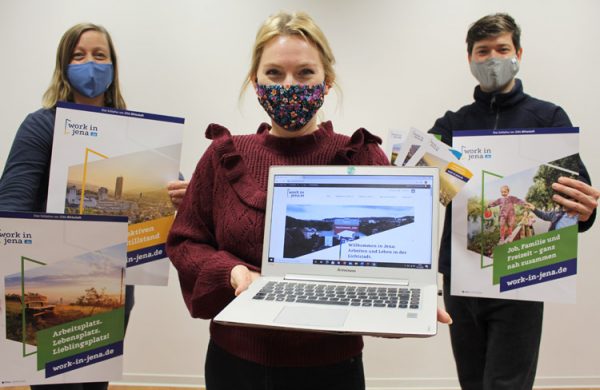 3 Personen mit Mund-Nasen-Bedeckung stehen nebeneinander, halten Plakate und Laptop in Kamera