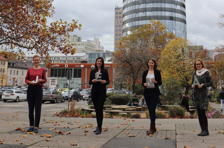 4 Frauen stehen nebeneinander auf großem Platz, halten Buchstaben in der Hand, im Hintergund Gebäude und Parkplatz