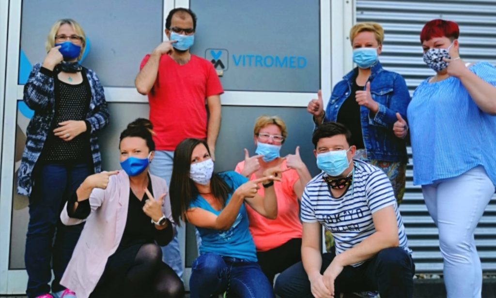 A group of men and women wearing mouth guards pose in front of the entrance door of the company VITROMED GmbH.
