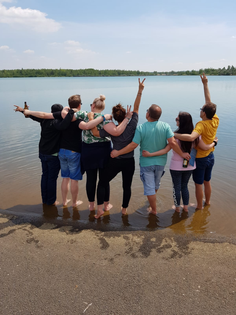 A group of men and women stand in front of a lake with their backs to the camera, holding each other in their arms and looking at the lake.