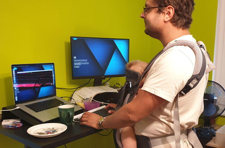 Mann steht vor Laptop, Tastatur und Bildschirm auf einem Schreibtisch, trägt Babytrage mit Kind vor der Brust