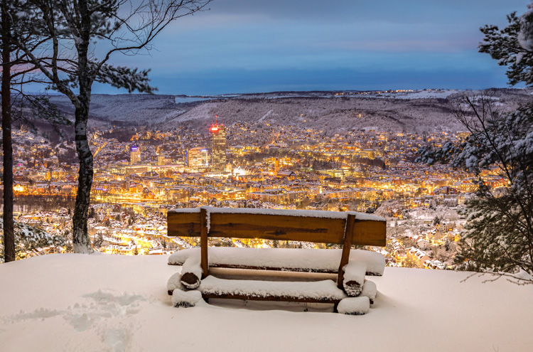 Im Vorderung Holzbank mit Schnee bedeckt, dahinter Blick auf eine Stadt bei Dämmerung, viele gelbe Lichter, im Hintergrund verschneite Landschaft