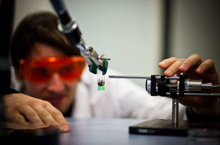 technische Aparatur, die ein Reagenzglas mit grüner Flüssigkeit hält, im Hintergrund Mann mit Kittel und oranger Brille