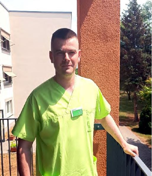 Profilfoto eines jungen Mannes in hellgrüner Krankenpfleger-Uniform am Balkon stehend.