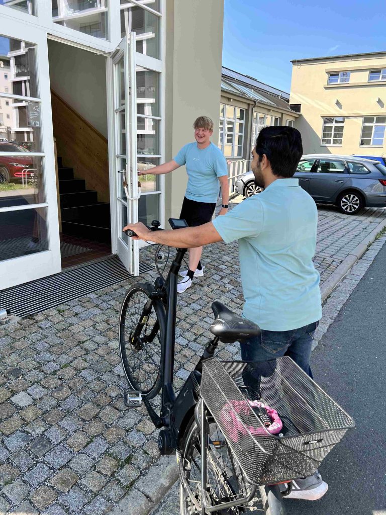 Eine Person öffnet einer anderen Person mit Fahrrad die Tür zu einem Gebäude.
