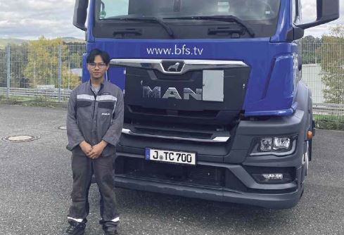 Young man in work clothes stands in front of a blue lorry.