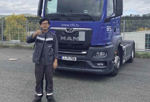 Young man in work clothes stands in front of a blue lorry and shows "thumbs up".
