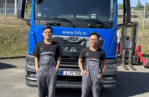 Two young men in work clothes stand in front of a blue lorry.