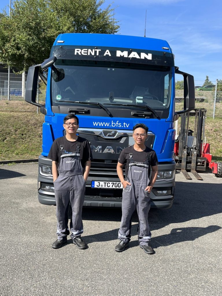 Two young men in work clothes stand in front of a blue lorry.